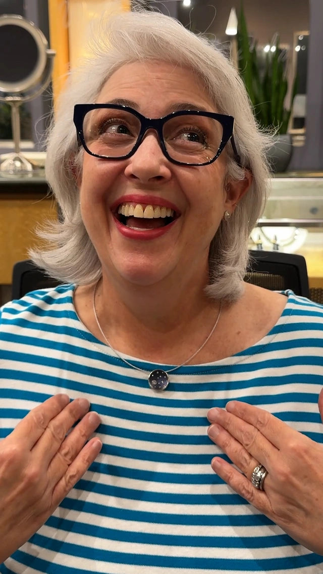 Woman with gray hair and glasses wearing a blue and white striped shirt, smiling showing off her new Jewelsmith custom pendant.